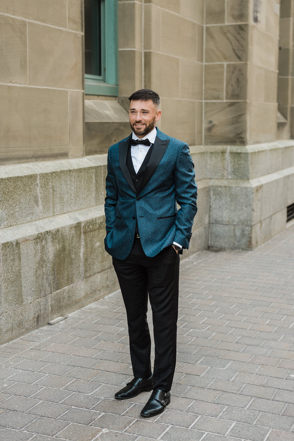 Stylish groom poses in front of beige stone building in downtown Halifax