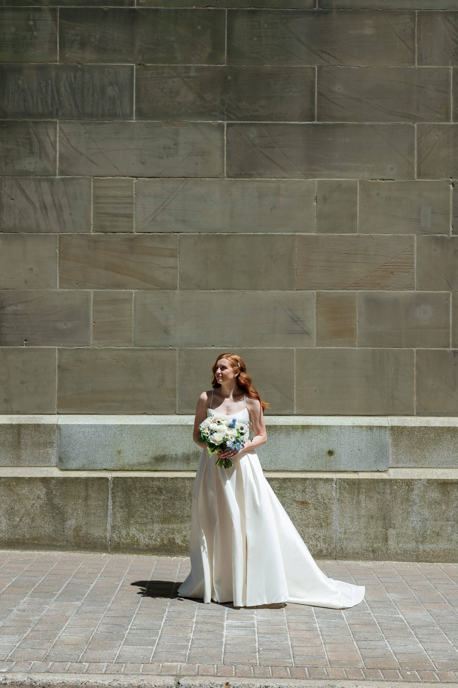 bride poses for portrait on halifax street. She has beautiful red hair and is holding blue and white flowers