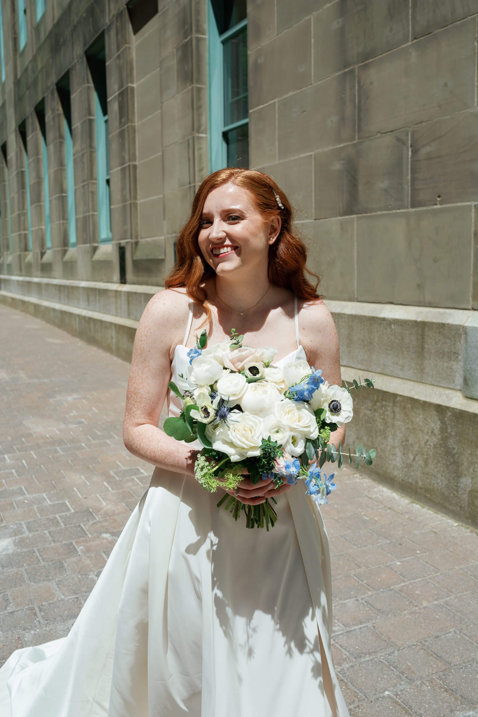 bride poses for portrait on halifax street. She has beautiful red hair and is holding blue and white flowers